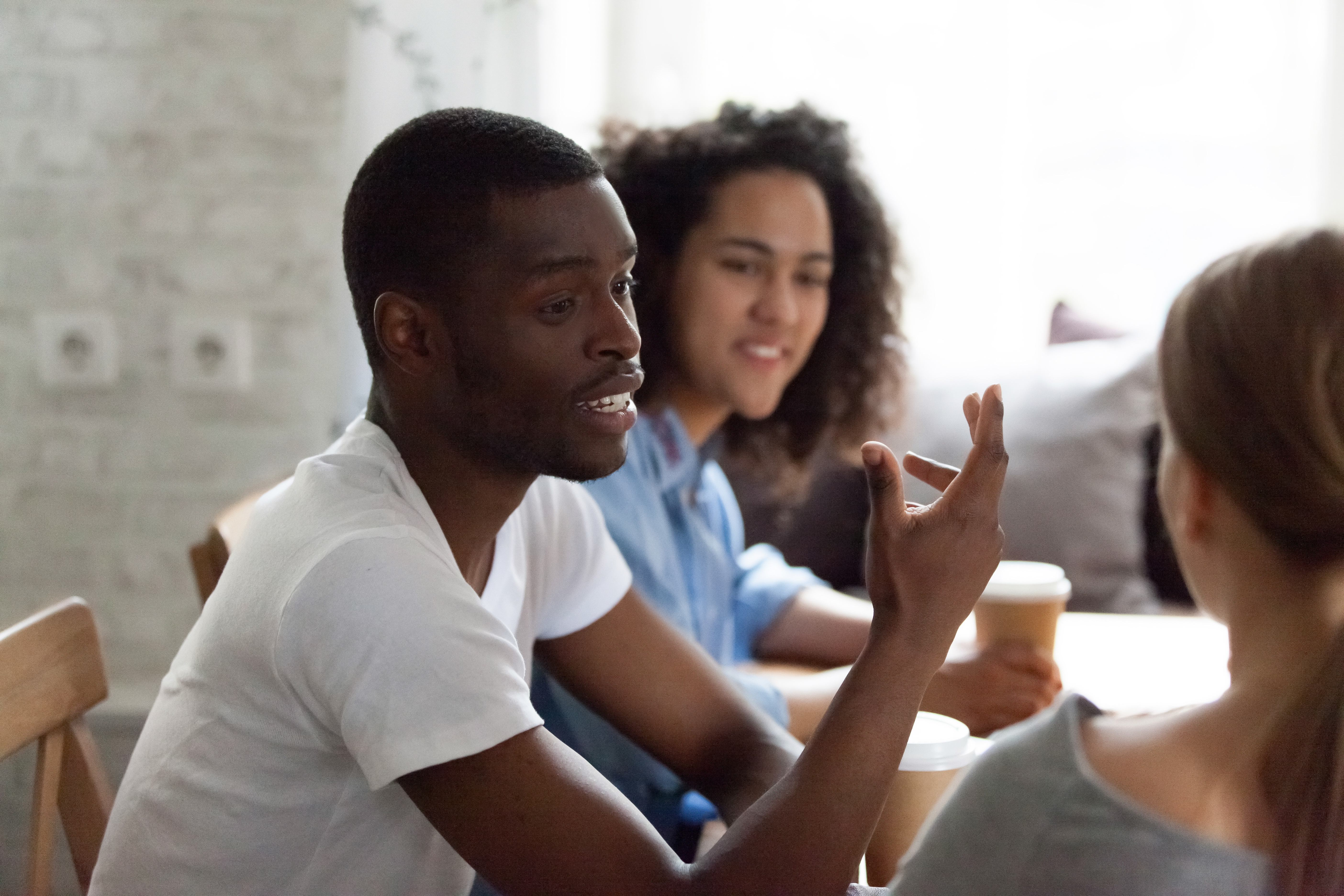 Smiling happy african american guy sitting at table in cafe, telling stories, gesticulating, diverse multiracial female friends listening, talking, enjoying free time with best friends or colleagues.